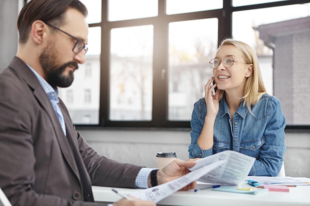 Portrait of serious bearded male model looks attentively at documents, studies terms of contract while female colleague speaks with financial partners, tries to solve all problems, answer questions